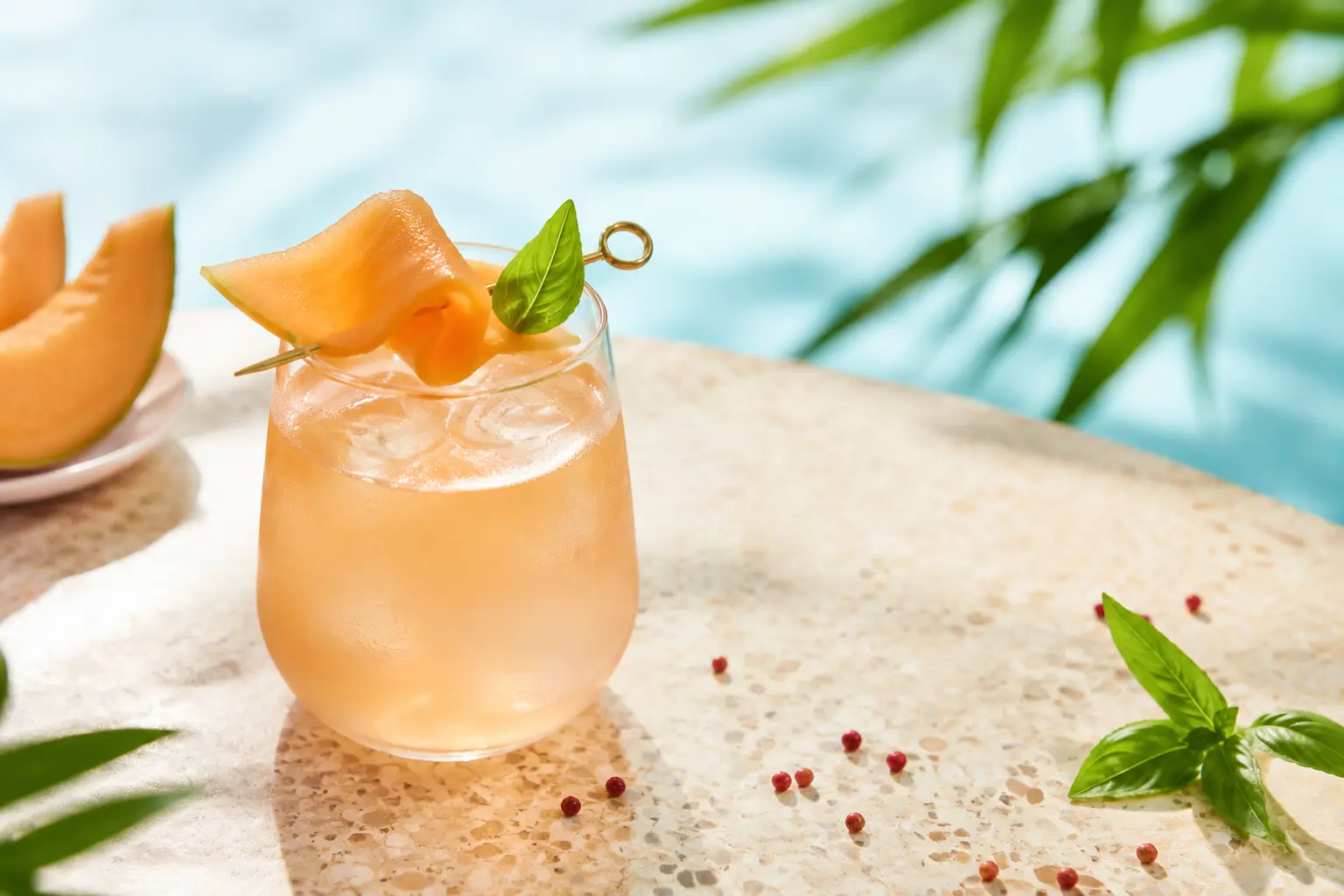 Pale coral alcohol-free cantaloupe and verjuice cooler over clear ice with a melon ribbon and basil garnish
